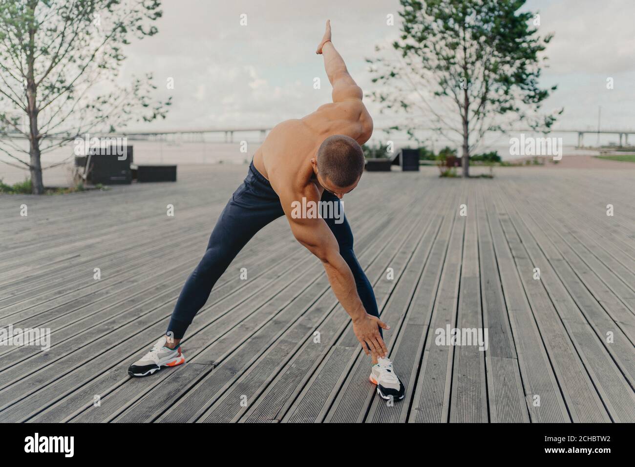 Beau sportif motivé non rasé fait des exercices sur la promenade, se réchauffe avant de courir, vêtu de vêtements actifs, pose à l'extérieur. Coureur mâle avec na Banque D'Images