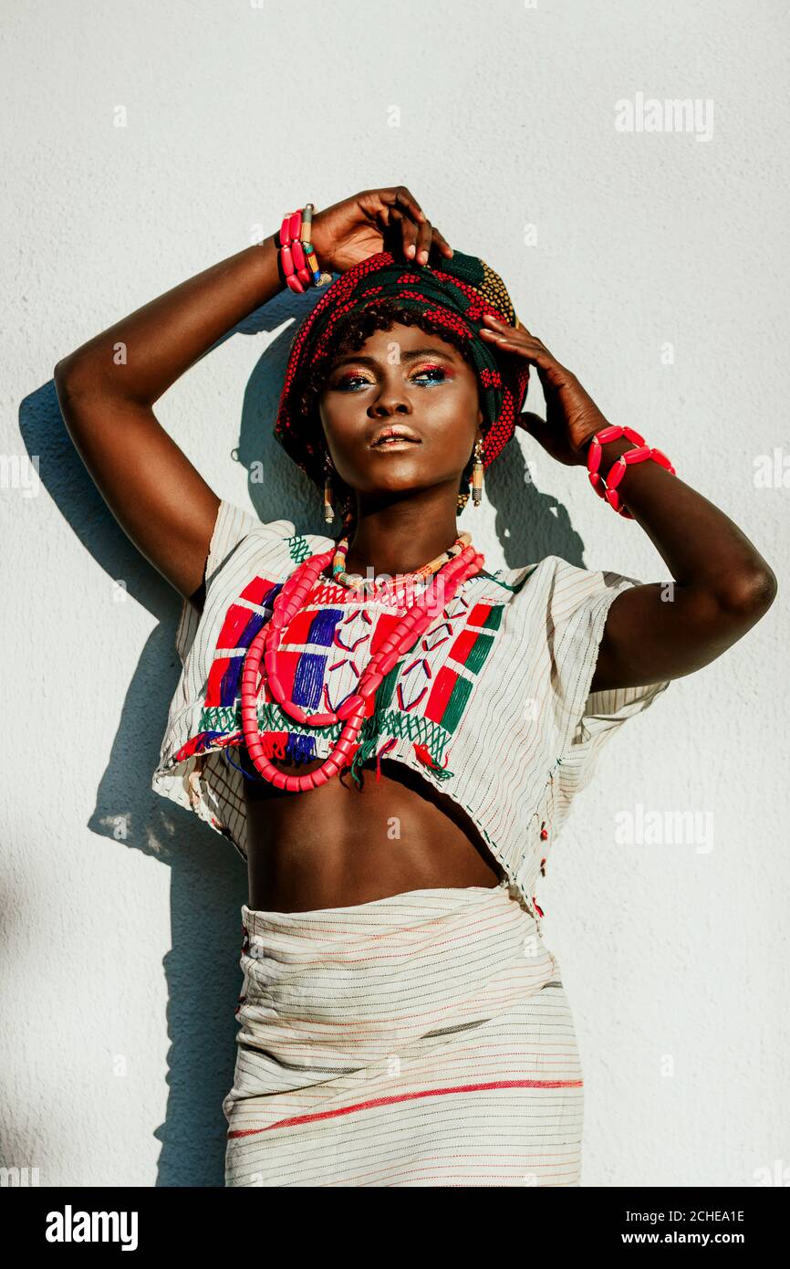 image rognée du modèle féminin africain de mode en costume national, bijoux, turban et maquillage posant sur fond de mur blanc, soleil de lumière du jour. Banque D'Images
