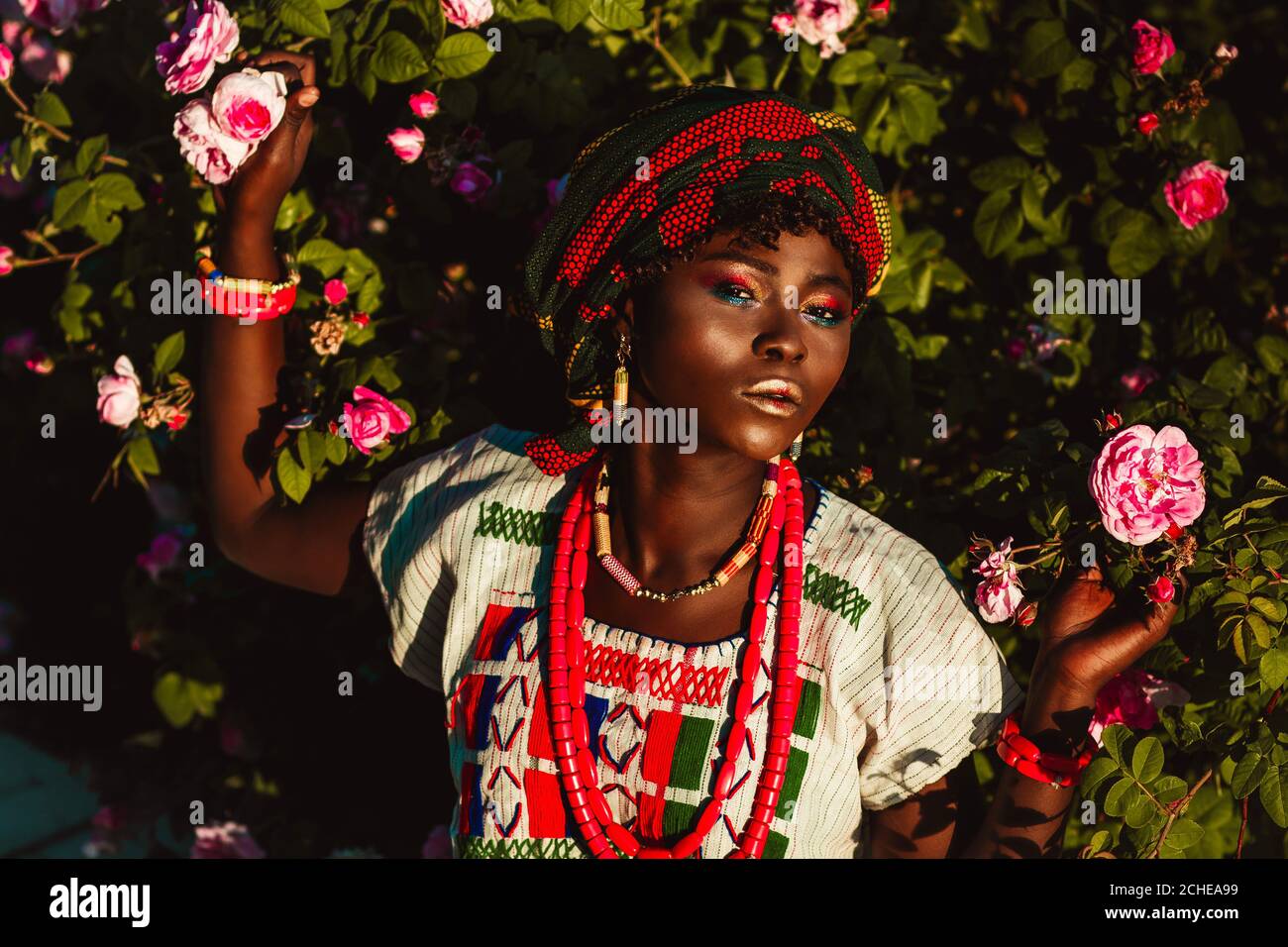 image rognée du modèle féminin africain de mode en costume national, bijoux, turban et maquillage posant dans le jardin de roses, lumière du jour lumineuse. Banque D'Images