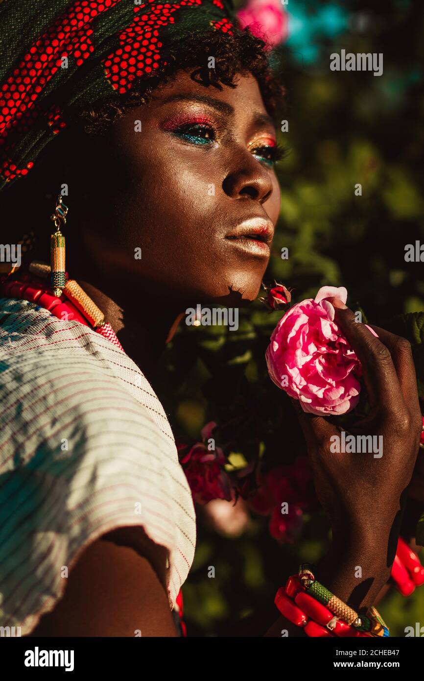 image rognée du modèle féminin africain de mode en costume national, bijoux, turban et maquillage posant dans le jardin de roses, lumière du jour lumineuse. Banque D'Images