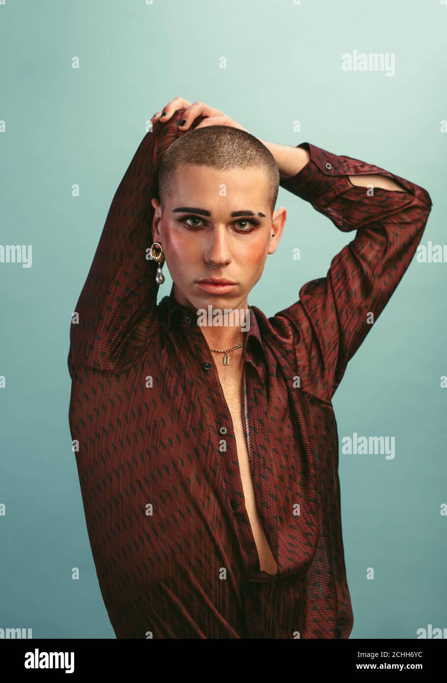 Homme gay avec maquillage portant des boucles d'oreilles debout au studio. Homme transgenre en chemise de soie posant sur fond bleu. Banque D'Images
