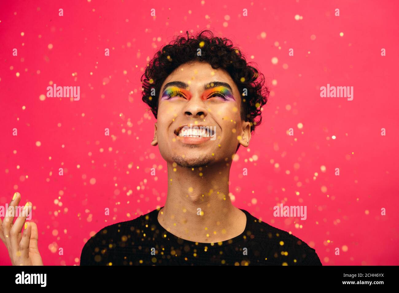 Homme plein de gaieté avec maquillage pour les yeux arc-en-ciel qui jette les paillettes. Excitée sexe liquide mâle regardant tomber paillettes sur fond rouge. Banque D'Images