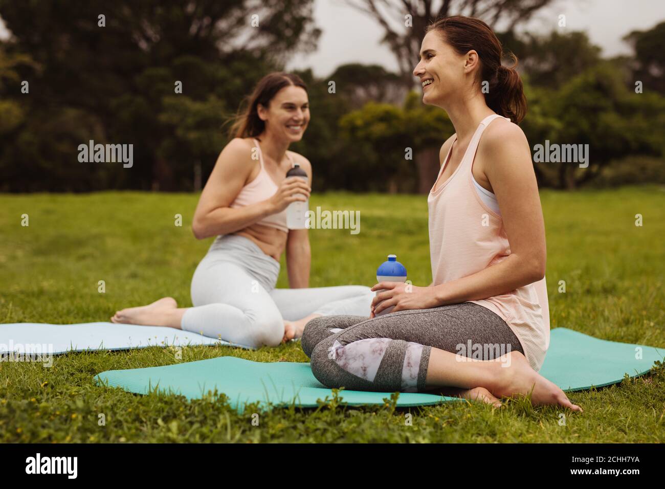 Des femmes gaies se détendent pendant une séance d'entraînement. Des femmes de fitness souriantes assises sur des tapis de yoga dans un parc d'eau potable. Banque D'Images