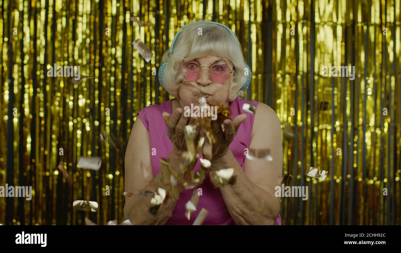 Bonne vieille femme âgée riant, soufflant confetti paillettes, célébrant anniversaire félicitant, gagnant loterie. Vieille dame élégante grand-mère sur fond doré Banque D'Images