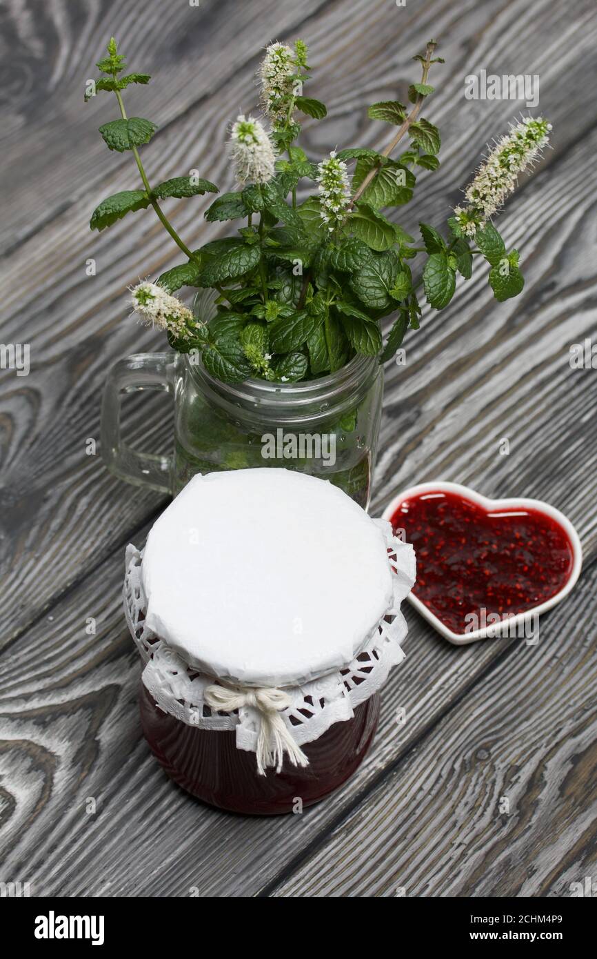 Confiture de framboises en pots et soucoupe. Sur les planches de pin peintes en noir et blanc. Branches fleuries de menthe à proximité dans un pot. Banque D'Images