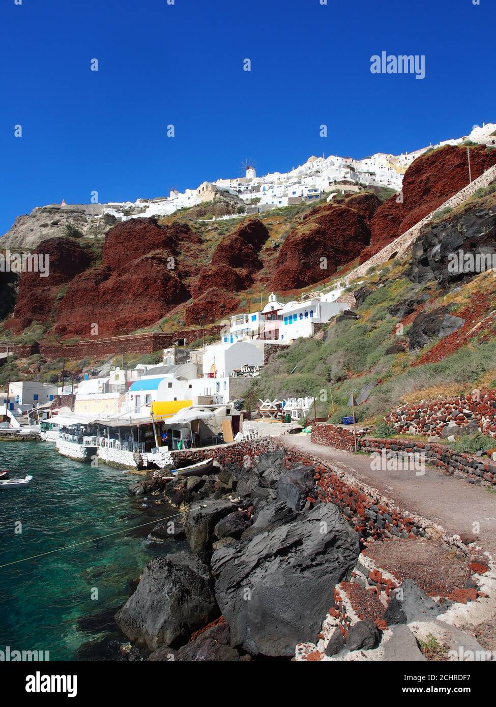 Des bateaux amarrés dans la baie d'Amoudi sous un ciel bleu clair et contre des falaises rouges et des maisons blanches d'Oia, Santorin Banque D'Images