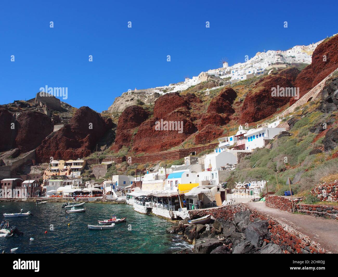 Des bateaux amarrés dans la baie d'Amoudi sous un ciel bleu clair et contre des falaises rouges et des maisons blanches d'Oia, Santorin Banque D'Images