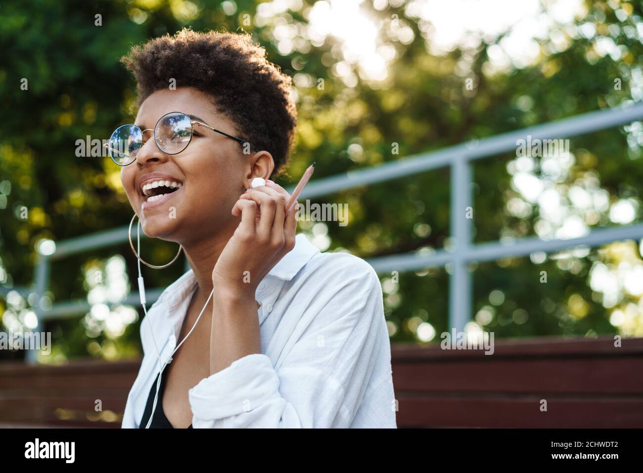 Photo d'une femme africaine optimiste en riant, assise à l'extérieur dans le parc tout en écoutant de la musique Banque D'Images