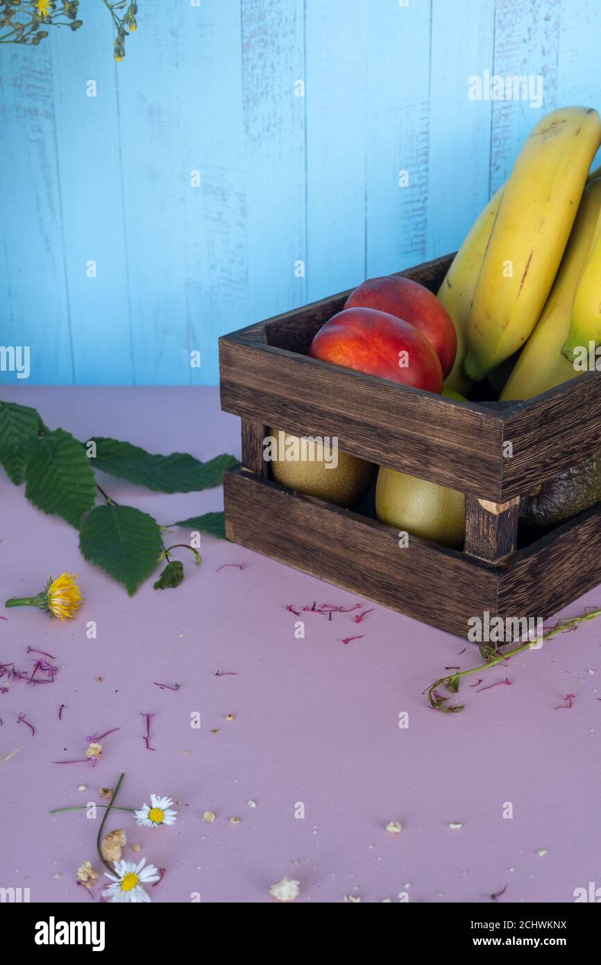 Boîte de fruits en bois avec banane, orange, kiwi, pêche, abricot et fleurs sur table rose avec fond bleu en bois Banque D'Images