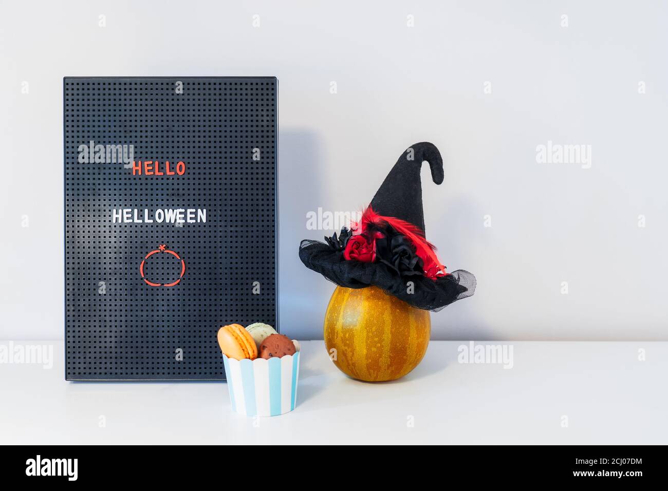 Décor minimaliste pour Halloween - panneau de lettres avec message Hello Halloween, chapeau de sorcière, citrouille décorative et tasse avec macarons doux sur fond blanc Banque D'Images