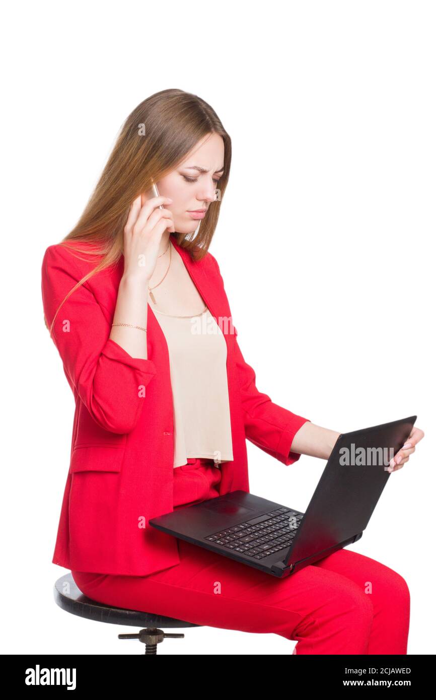 Une jeune femme de bureau d'affaires dans un costume rouge un arrière-plan isolé en blanc, tenant un ordinateur portable, et parlant sur un smartphone Banque D'Images