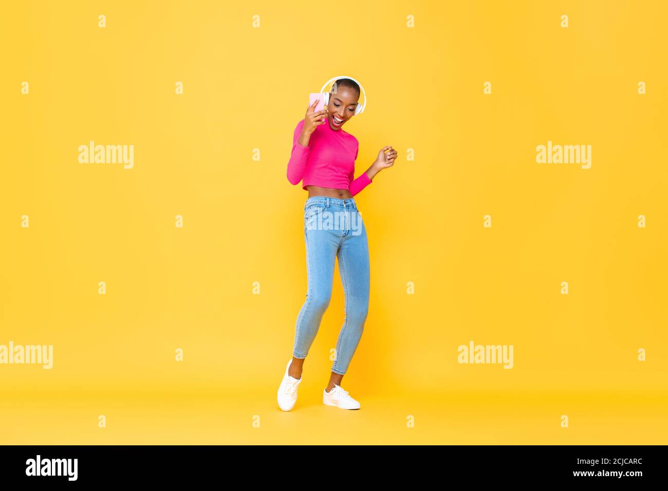 Portrait joyeux de la danse heureuse femme afro-américaine portant des écouteurs écouter de la musique depuis un smartphone sur un studio jaune coloré isolé arrière Banque D'Images