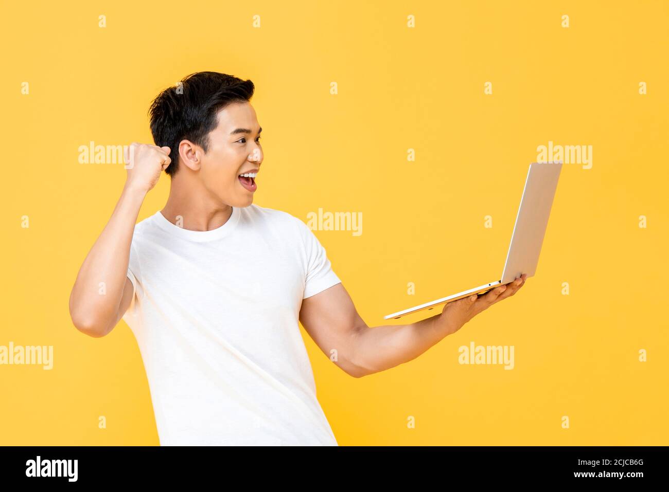 Un jeune homme asiatique enthousiaste qui regarde un ordinateur portable et se relève son poing faisant le geste oui isolé sur fond jaune Banque D'Images