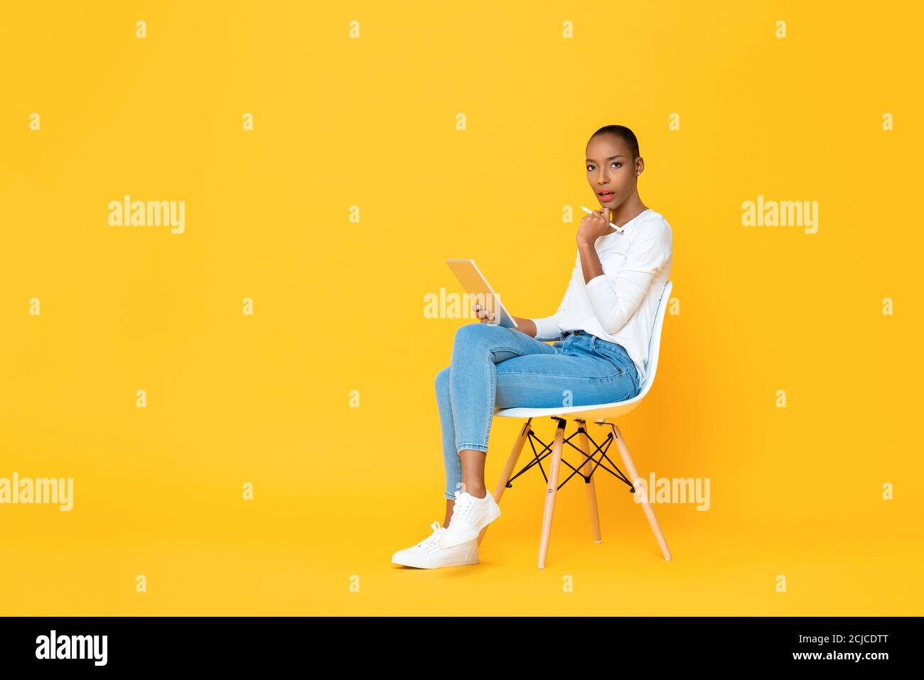 Penser jeune femme afro-américaine assis et utilisant un ordinateur tablette avec stylet sur fond jaune isolé Banque D'Images