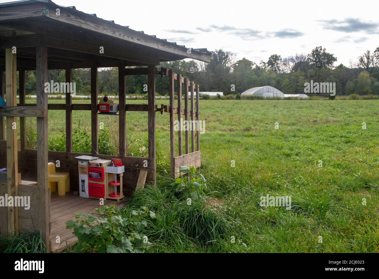 Un pavillon rustique avec des jouets se trouve dans un champ agricole vert sur une ferme familiale biologique avec un paysage rural et des serres en arrière-plan. Banque D'Images