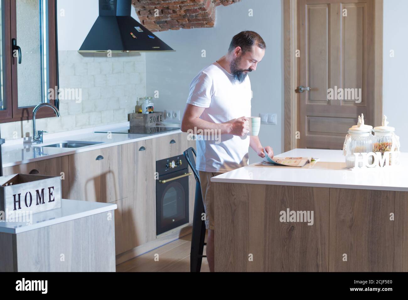 un homme de taille basse avec une barbe lisant un magazine tout en buvant café dans sa cuisine Banque D'Images