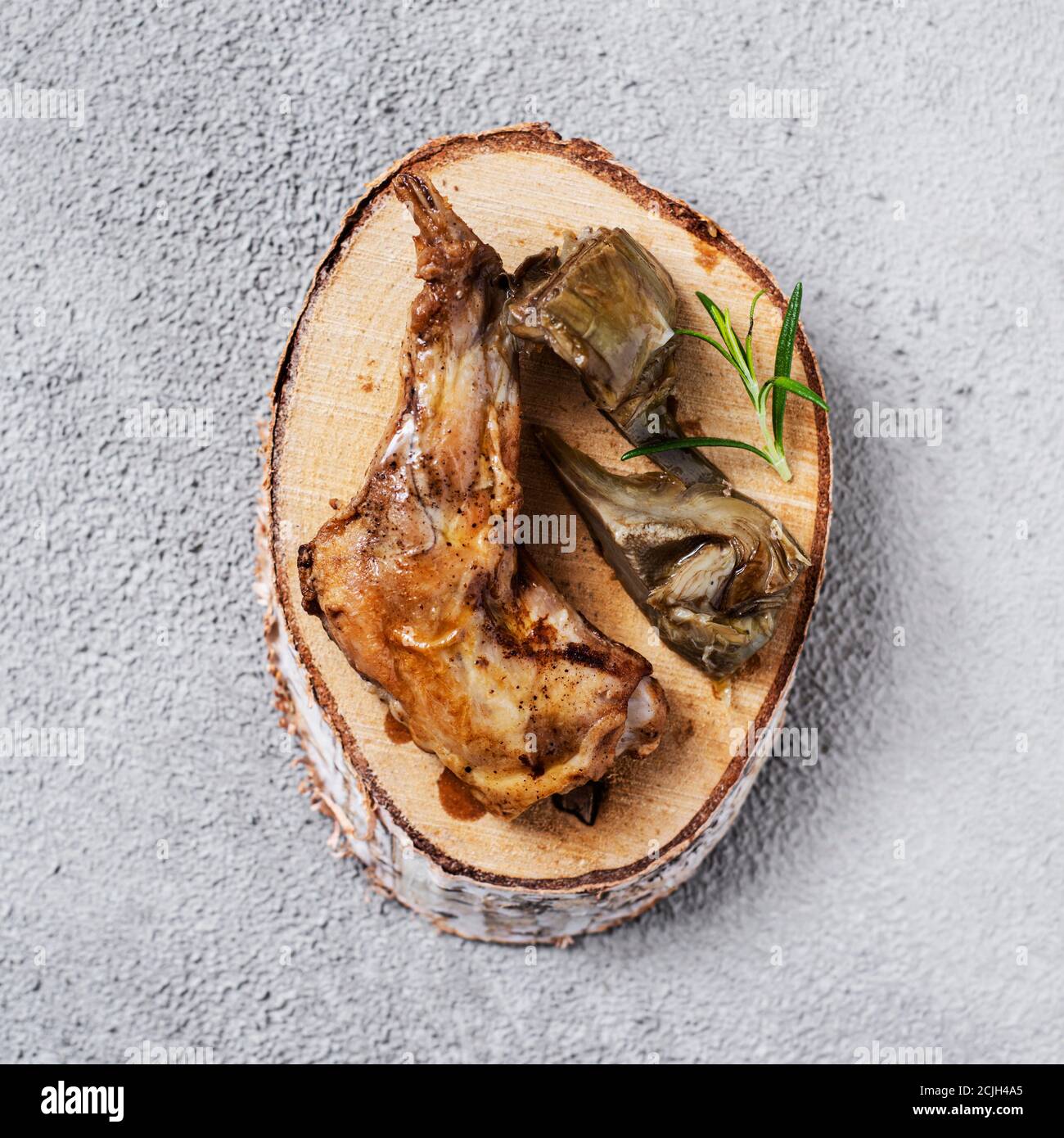 Vue en grand angle d'un morceau de lapin cuit, typiquement mangé en Espagne, et deux tranches d'artichaut sur un plateau en bois placé sur un béton texturé gris Banque D'Images