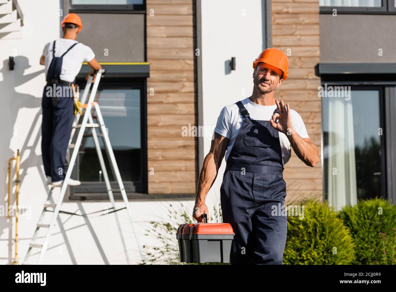 Mise au point sélective du constructeur en uniforme avec boîte à outils indiquant ok faites un geste pendant que votre collègue travaille près du bâtiment Banque D'Images