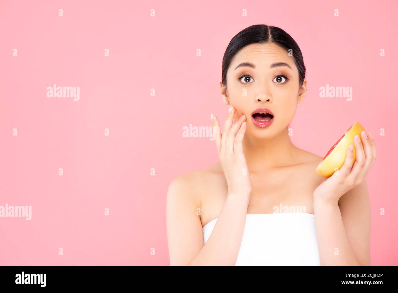 choqué femme asiatique touchant le visage tout en tenant la moitié de pamplemousse isolé sur rose Banque D'Images