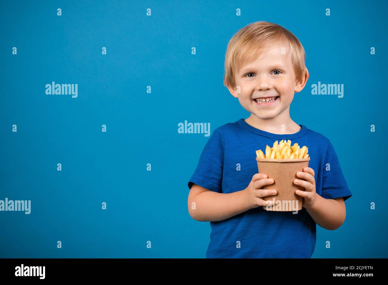 Garçon blond enfant de 4 ans heureux de tenir une tasse avec des frites dans ses mains. Gros plan studio tourné sur fond bleu avec espace pour le texte et la publicité f Banque D'Images