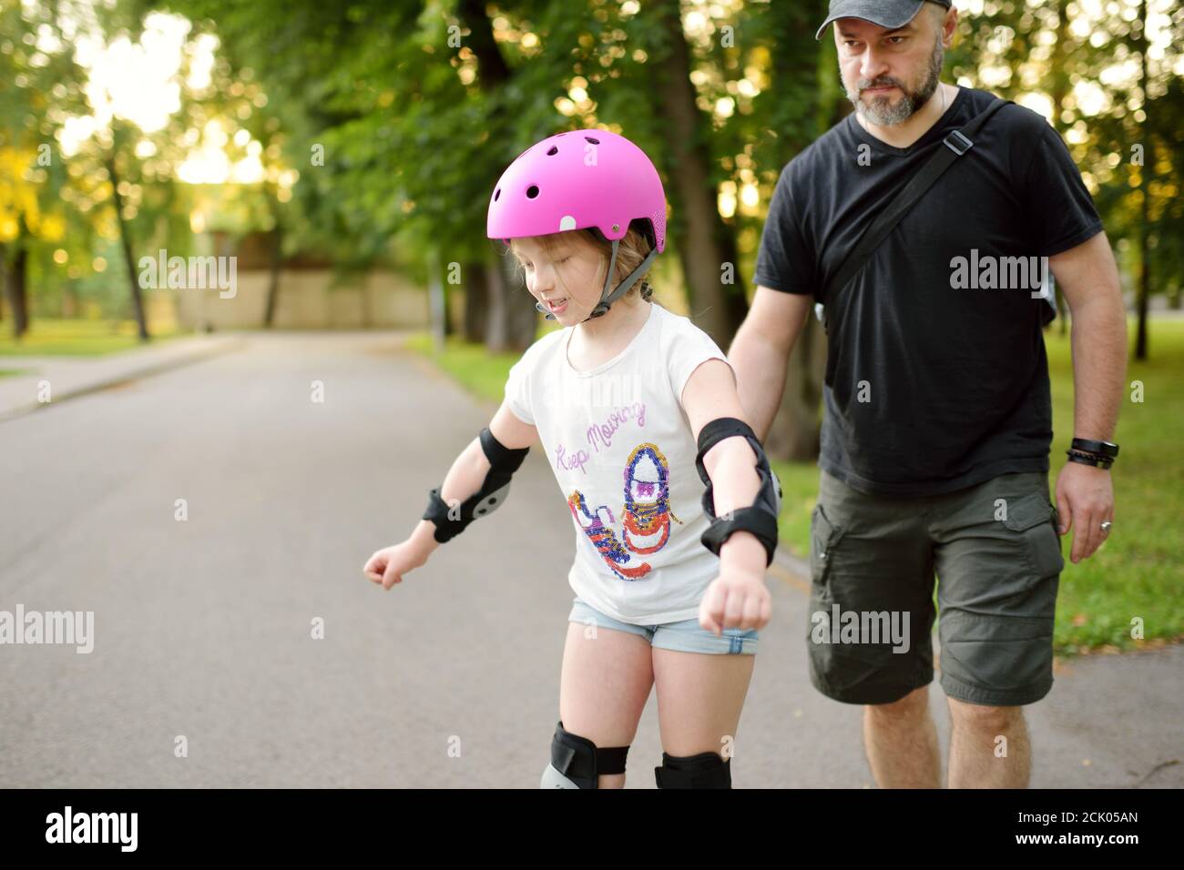 Père enseignant à sa jeune fille de skate à roulettes lors d'une belle journée d'été dans un parc. Enfant portant un casque de sécurité en train de patiner à roulettes à l'extérieur Banque D'Images