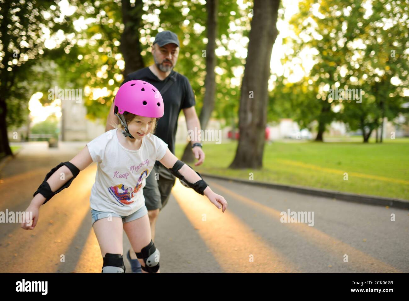 Père enseignant à sa jeune fille de skate à roulettes lors d'une belle journée d'été dans un parc. Enfant portant un casque de sécurité en train de patiner à roulettes à l'extérieur Banque D'Images