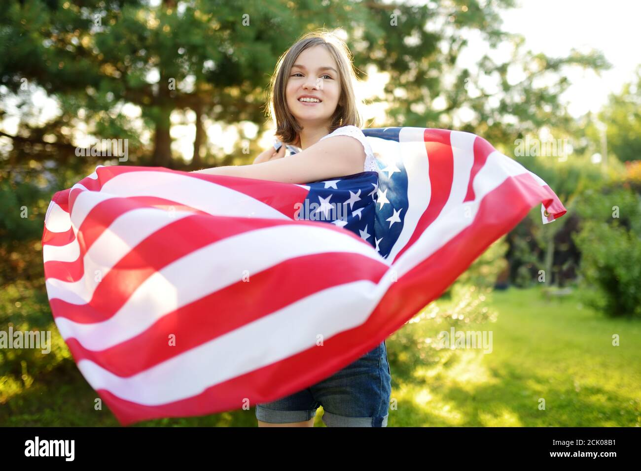 Jolie jeune fille portant un drapeau américain à l'extérieur le beau jour d'été. Concept de jour de l'indépendance. Enfant célébrant la fête nationale. Jour du souvenir. Banque D'Images