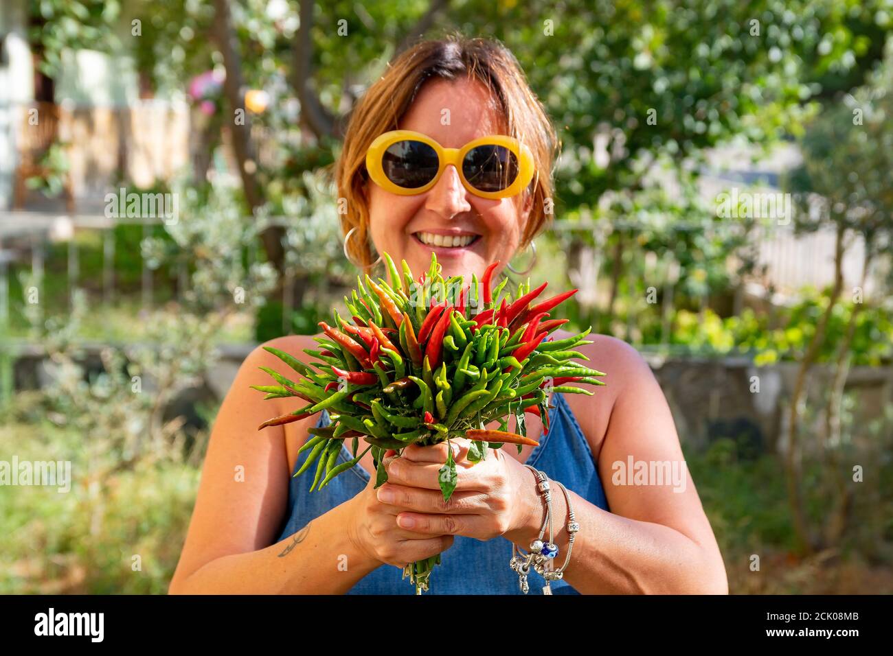 Femme brune caucasienne avec des lunettes de soleil jaunes tendance est tenue un bouquet de piments rouges et verts fraîchement récoltés Banque D'Images