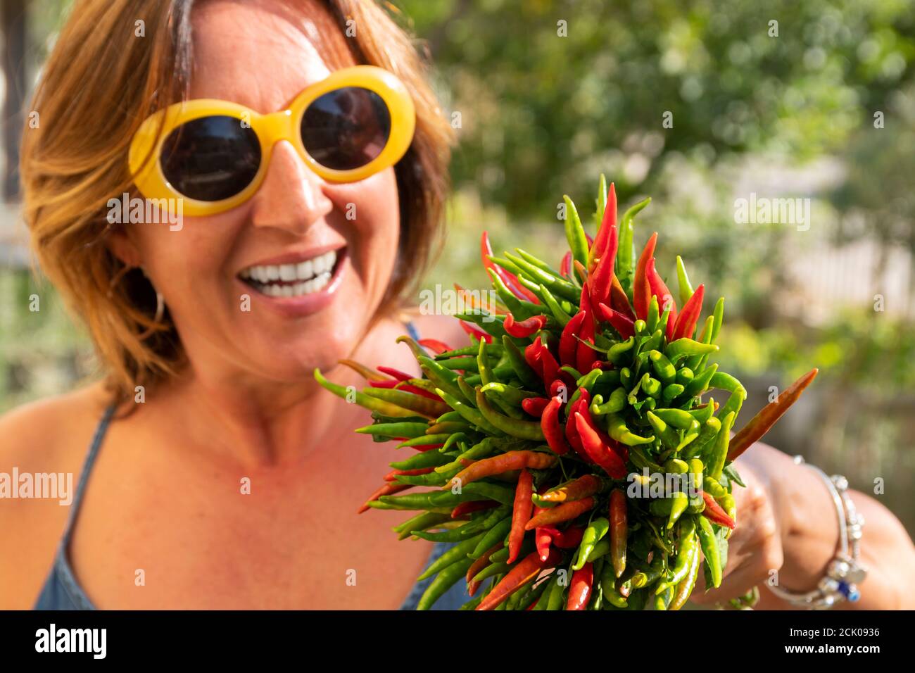 Gros plan sur un paquet de piments verts et rouges frais tenir par une belle femme de brunette caucasienne riante avec des lunettes de soleil jaunes élégantes. Banque D'Images