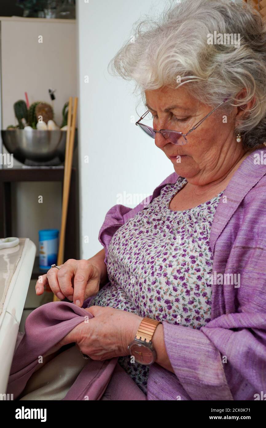 Une femme âgée aux cheveux gris est en cours de couture à la main à sa  maison. Elle porte des lunettes et une robe à fleurs violettes Photo Stock  - Alamy