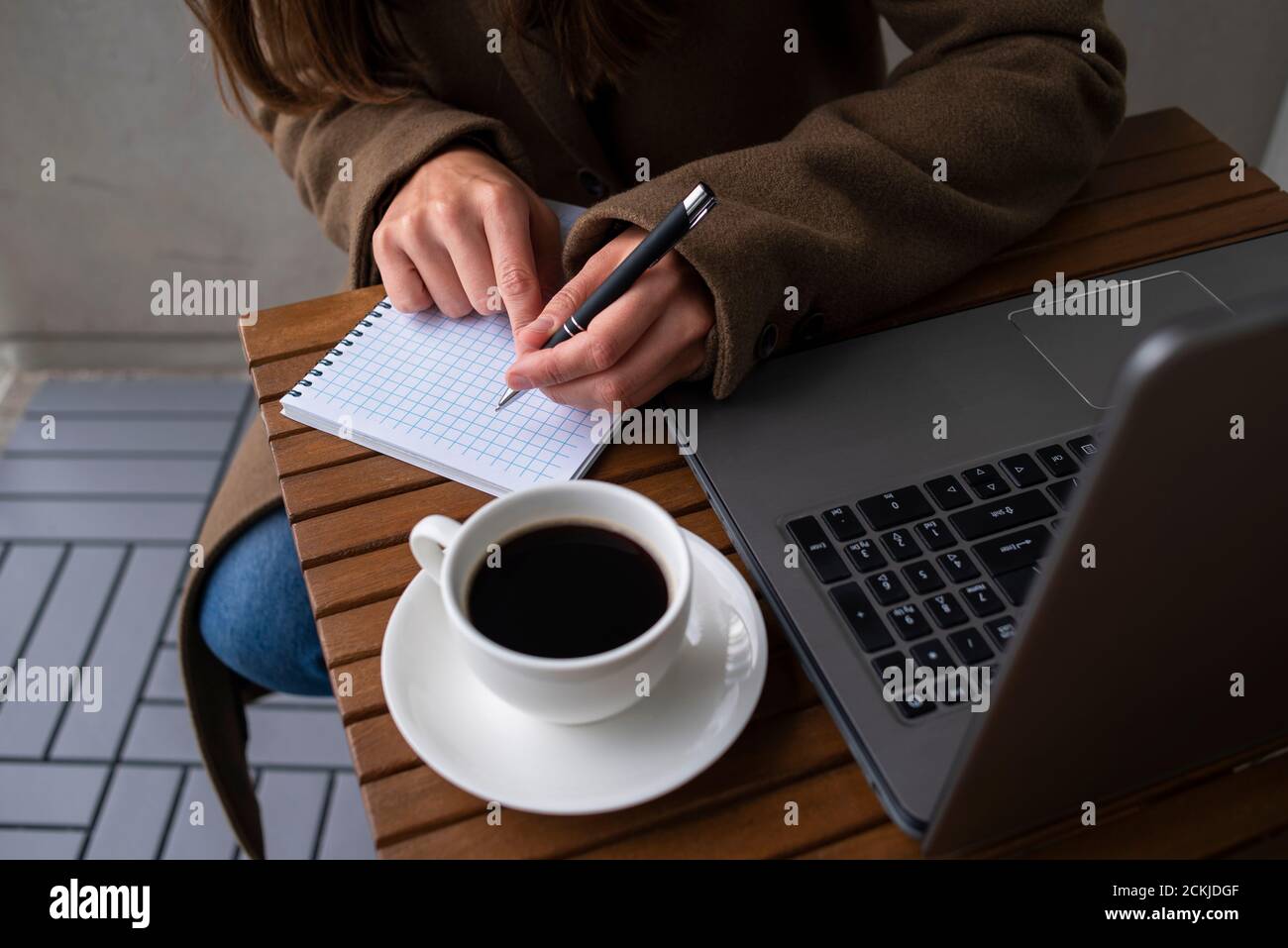 Femme en laine manteau travaillant dans un café de rue avec ordinateur portable. Une tasse de café sur une table en bois. Écriture dans le bloc-notes Banque D'Images