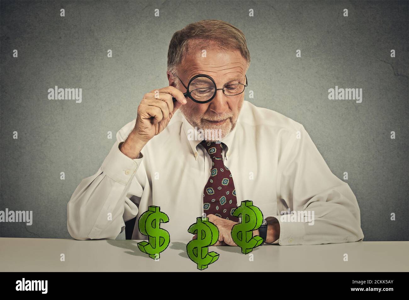 pincher penny. Homme d'affaires senior regardant à travers la loupe au dollar signe symbole sur la table isolé mur gris arrière-plan. Économie financière W Banque D'Images