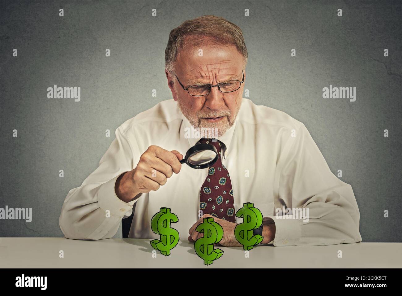 pincher penny. Homme d'affaires sceptique senior regardant à travers la loupe à dollar signe symbole isolé fond gris. Économie richesse financière Banque D'Images