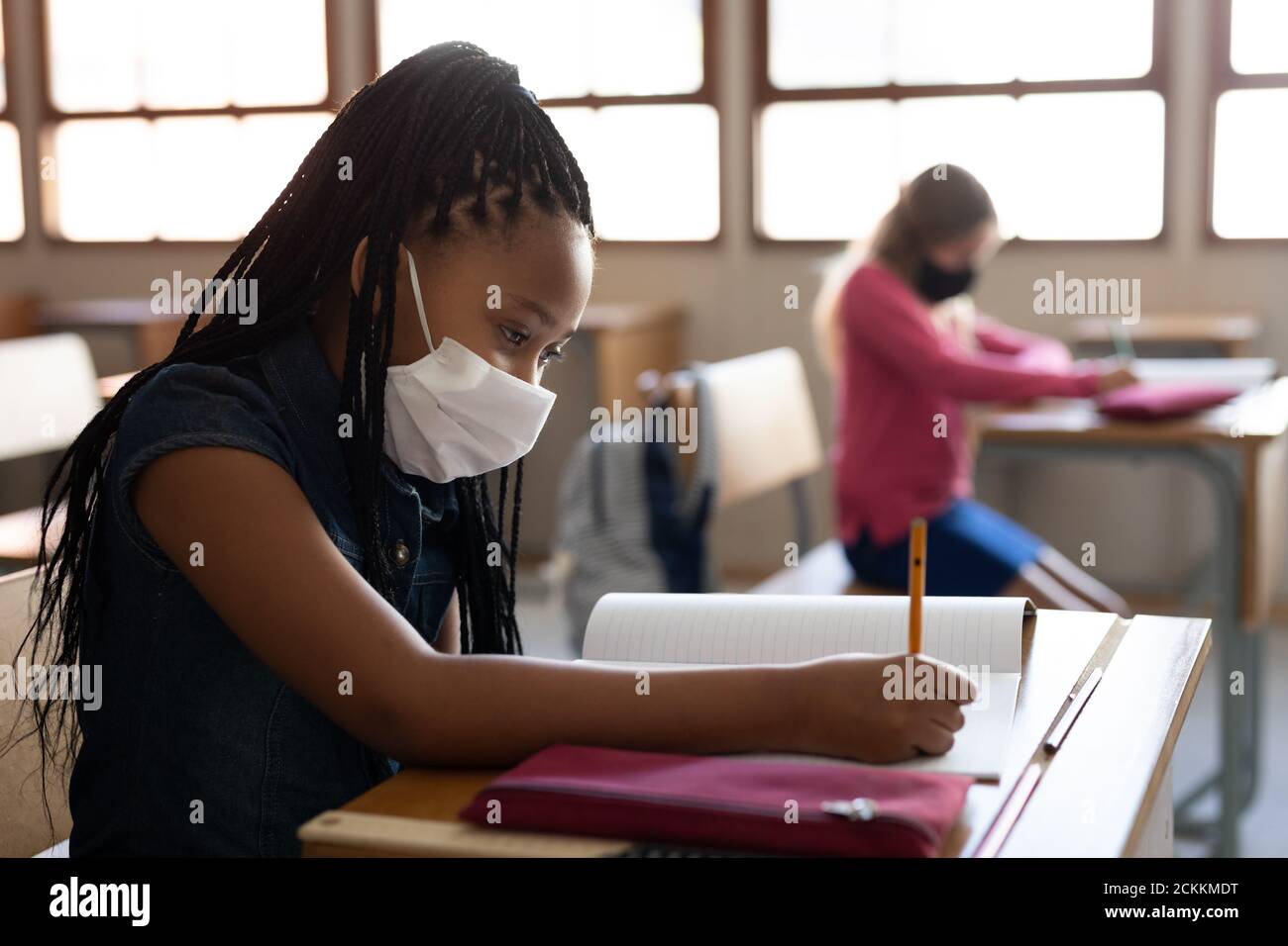 Fille portant un masque de visage écrivant tout en étant assise sur son bureau à l'école Banque D'Images
