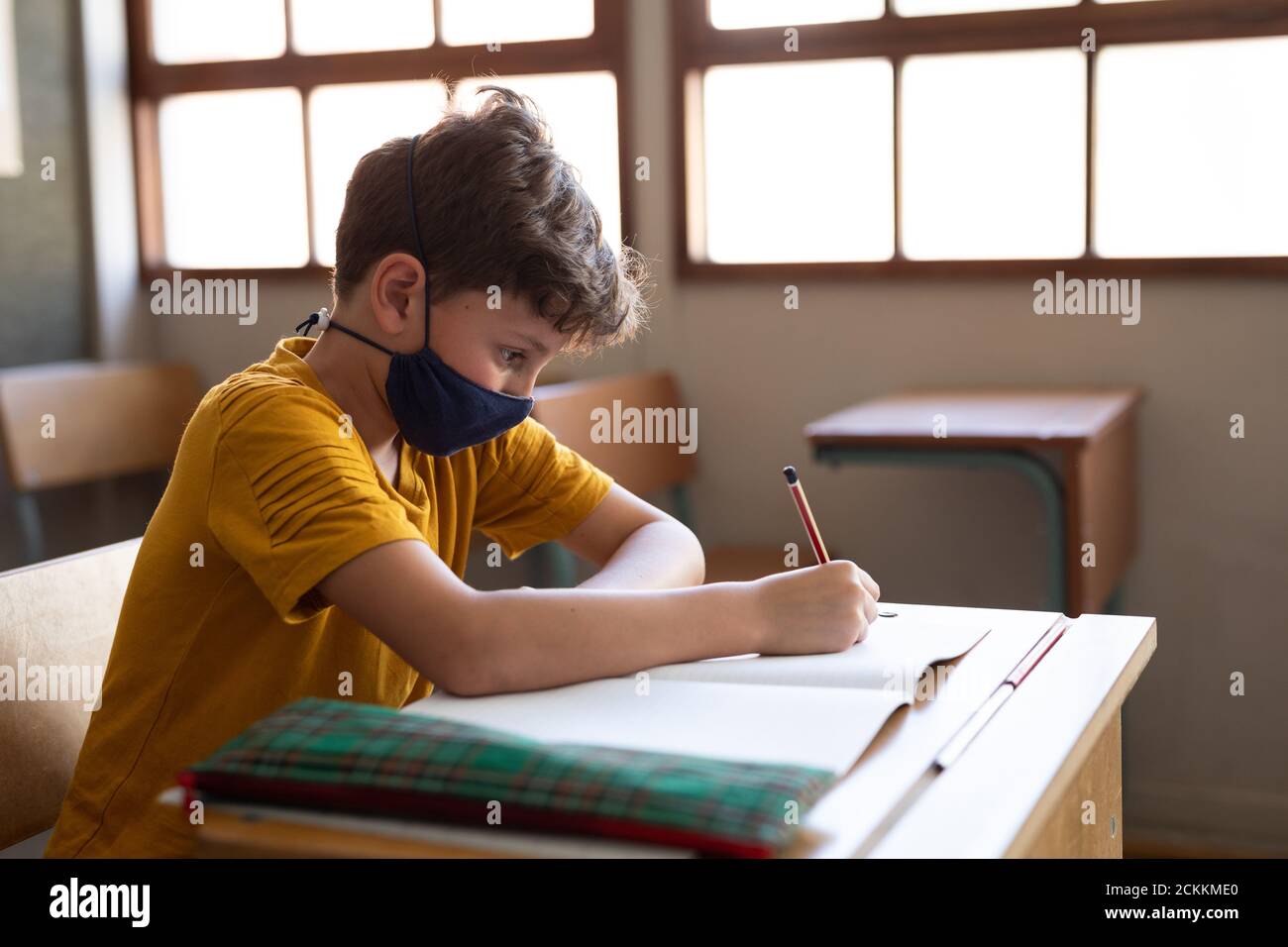 Garçon portant un masque de visage écrivant tout en étant assis sur son bureau à l'école Banque D'Images