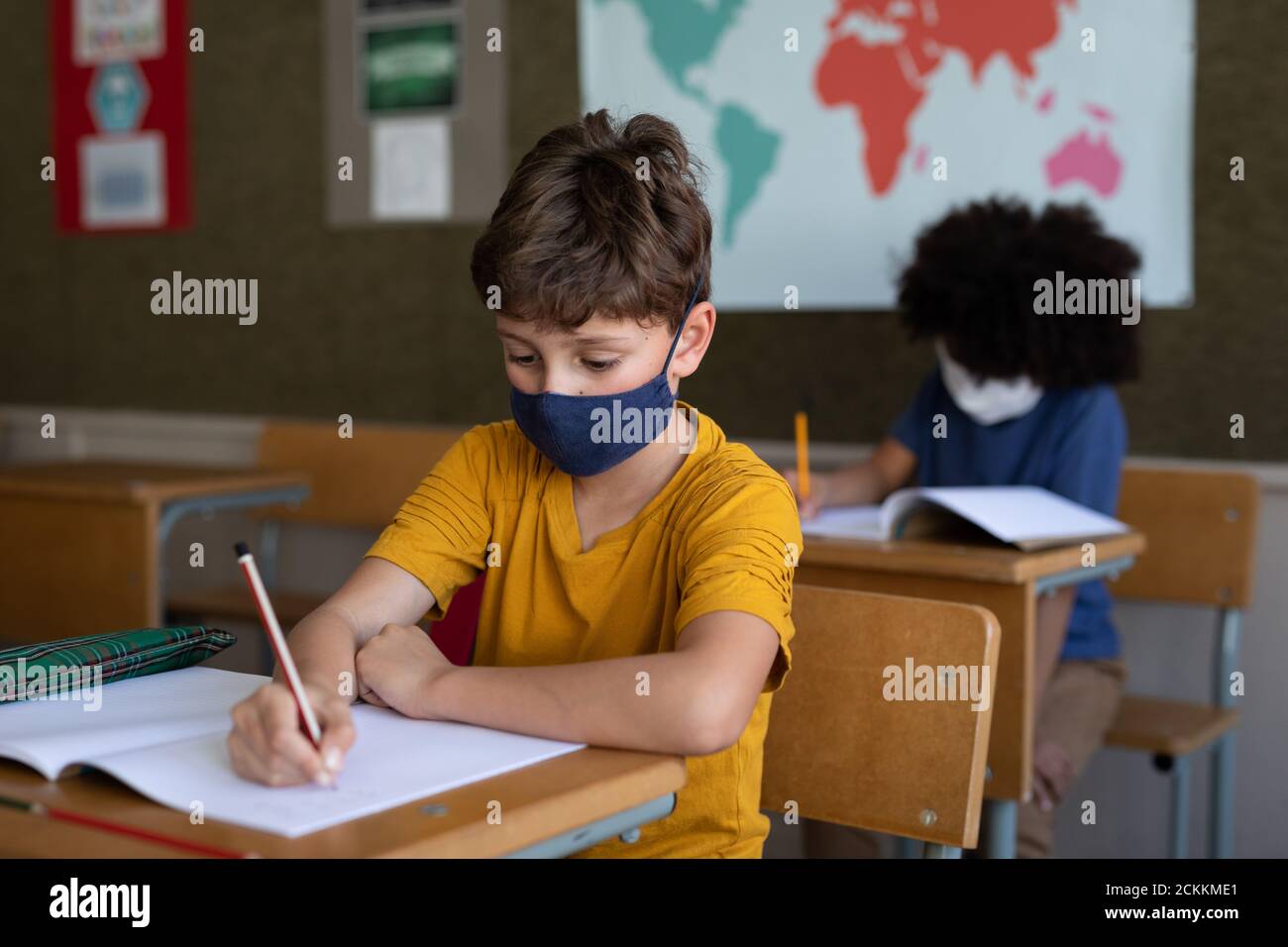 Garçon portant un masque de visage écrivant tout en étant assis sur son bureau à l'école Banque D'Images