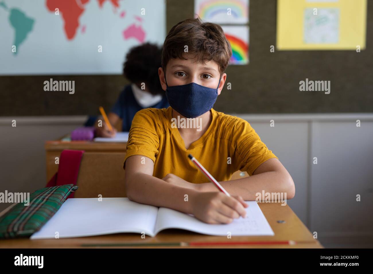 Portrait d'un garçon portant un masque de visage écrivant pendant qu'il est assis sur son bureau à l'école Banque D'Images