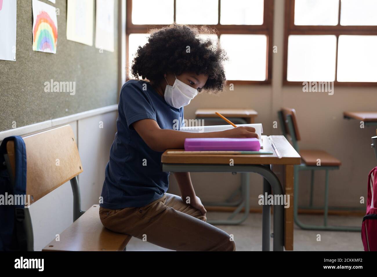 Garçon portant un masque de visage écrivant tout en étant assis sur son bureau à l'école Banque D'Images