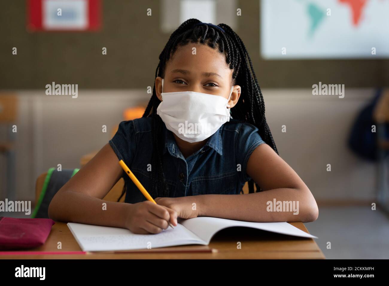 Portrait d'une fille portant un masque de visage écrivant pendant qu'elle est assise sur son bureau à l'école Banque D'Images