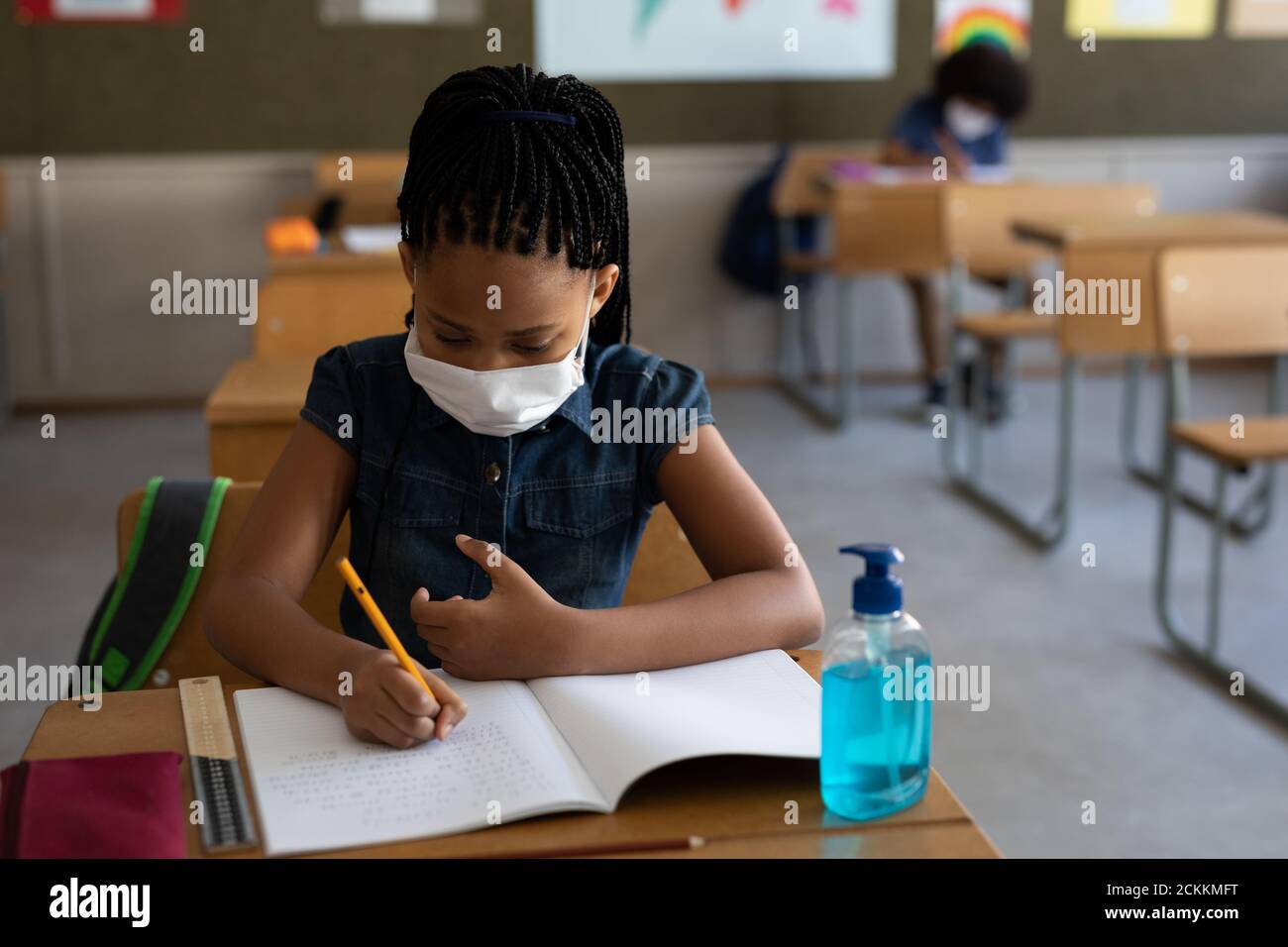 Fille portant un masque de visage écrivant tout en étant assise sur son bureau à l'école Banque D'Images