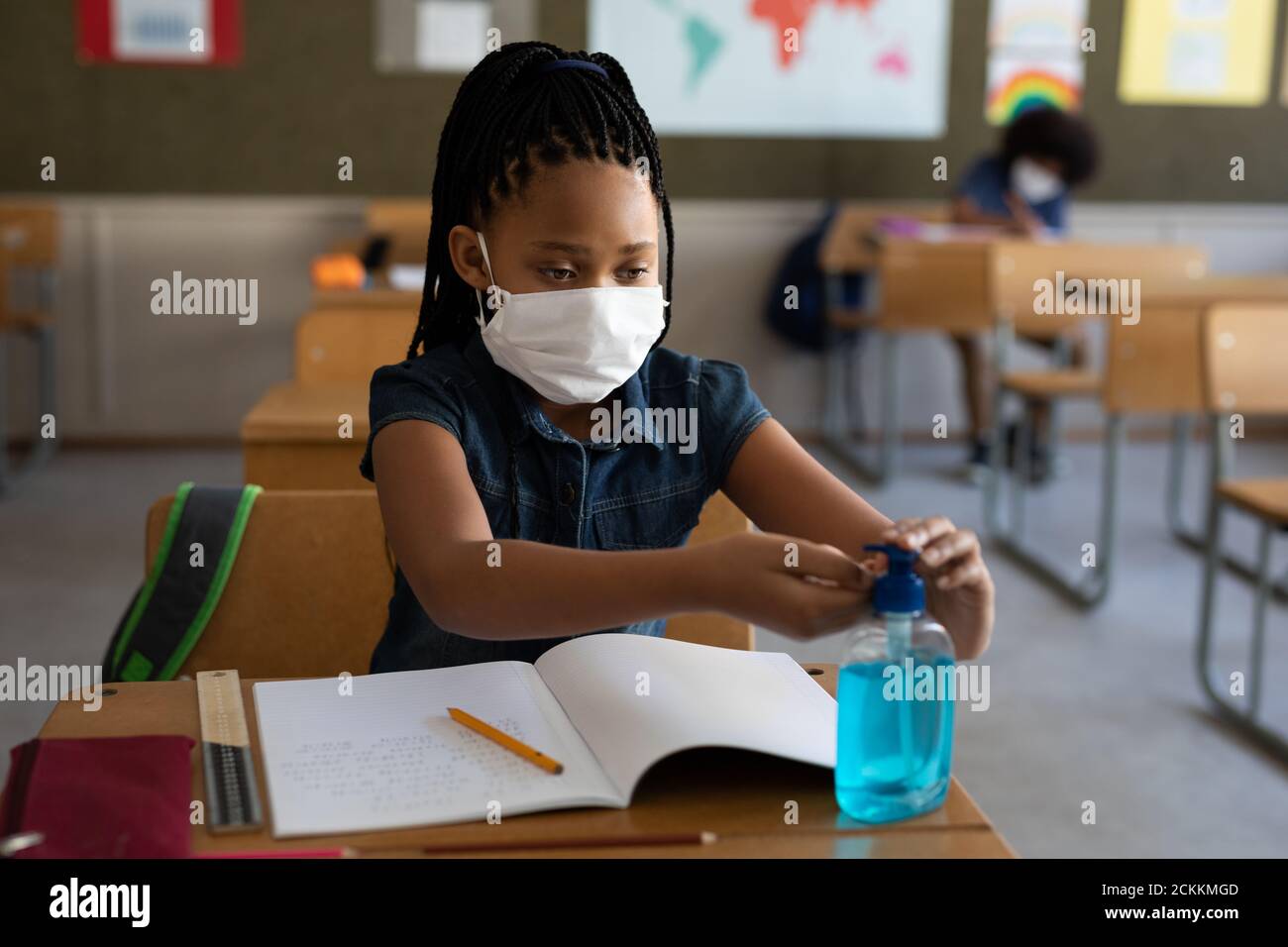 Fille portant un masque facial désinfectant ses mains tout en étant assise sur son bureau à l'école Banque D'Images