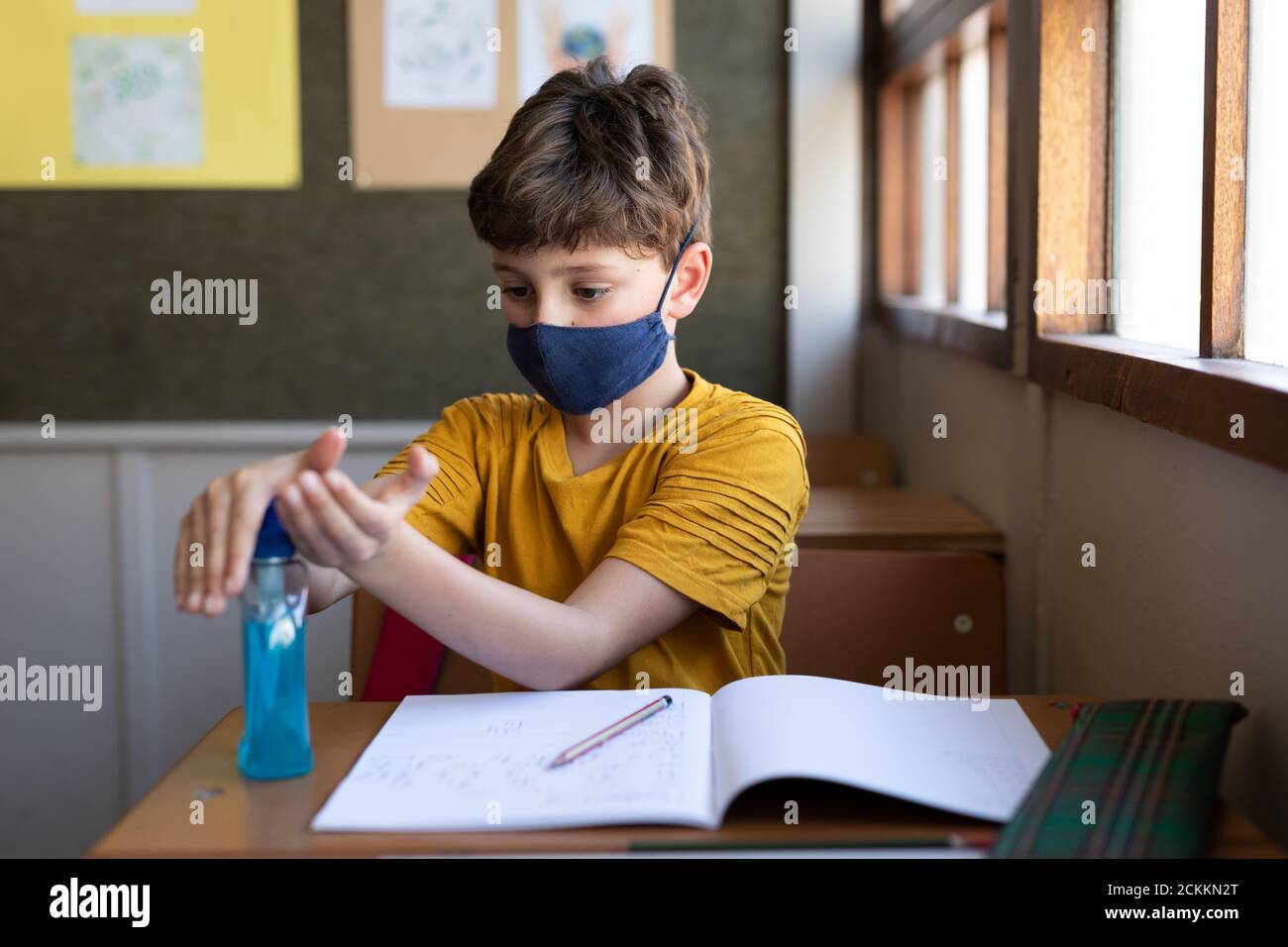 Garçon portant un masque désinfectant pour les mains tout en étant assis son bureau à l'école Banque D'Images