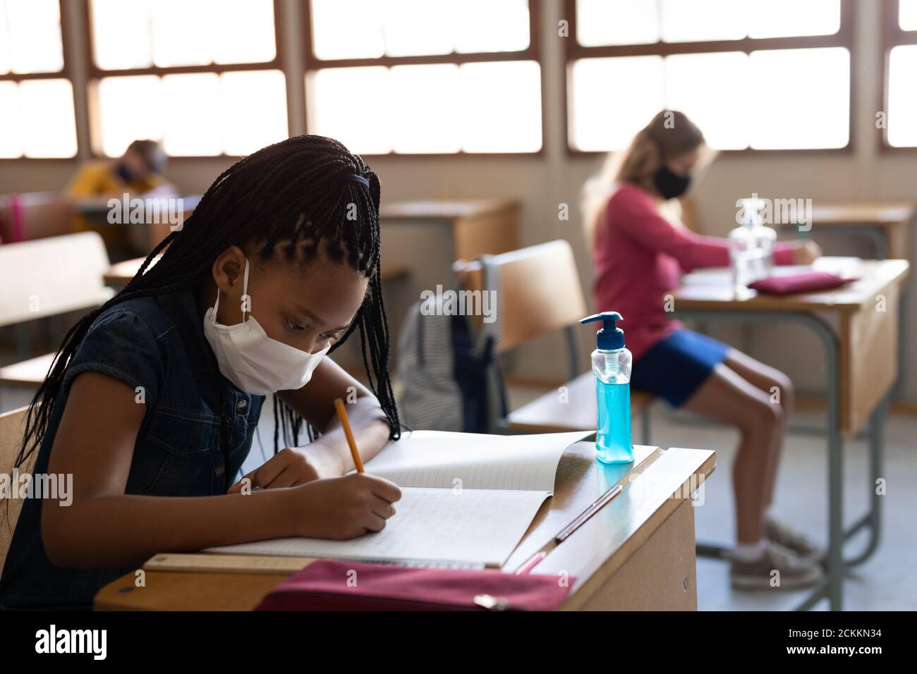 Fille portant un masque de visage écrivant tout en étant assise sur son bureau à l'école Banque D'Images