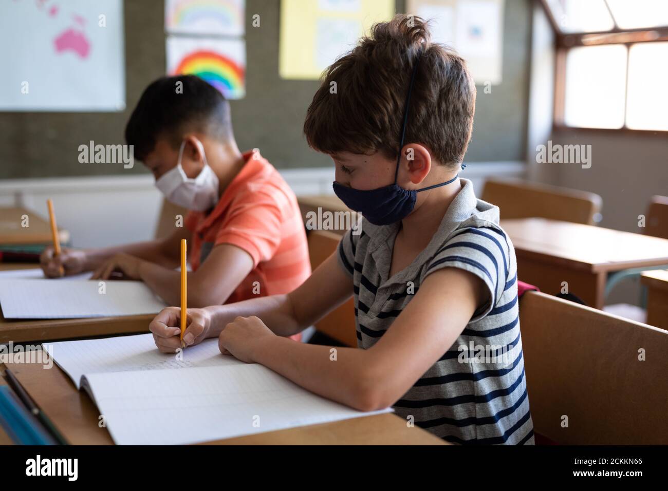 Garçon portant un masque de visage écrivant tout en étant assis sur son bureau à l'école Banque D'Images