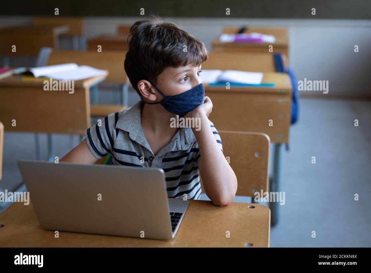 Garçon portant un masque facial assis avec un ordinateur portable sur son bureau à l'école Banque D'Images