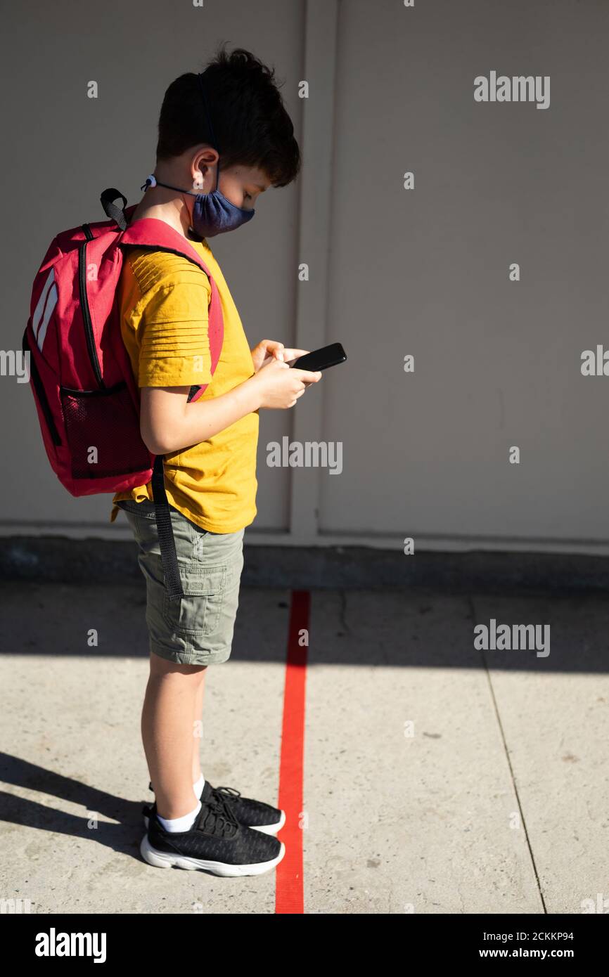 Garçon portant un masque facial avec un smartphone à l'école Banque D'Images