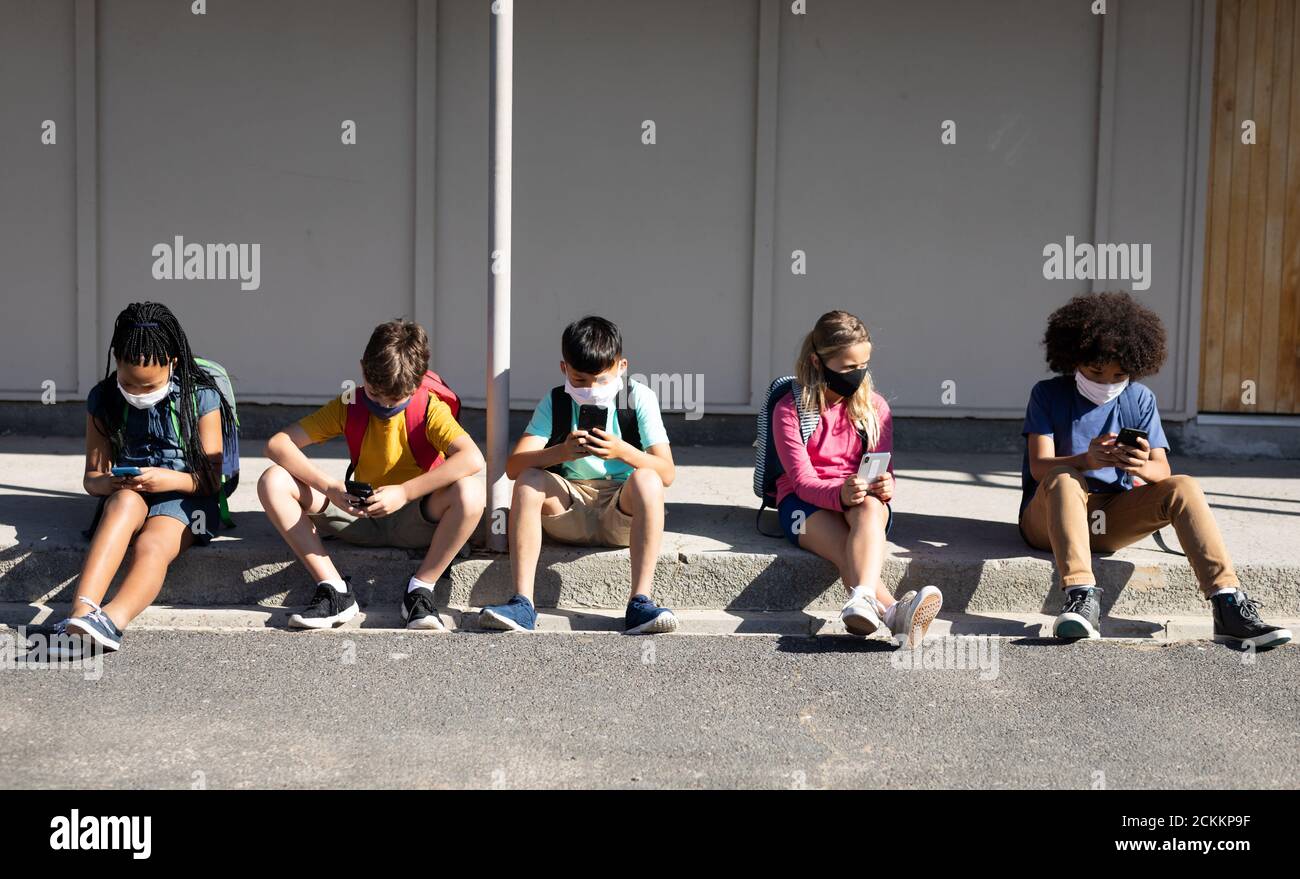 Groupe d'enfants portant un masque facial avec un smartphone lorsqu'ils sont assis ensemble Banque D'Images