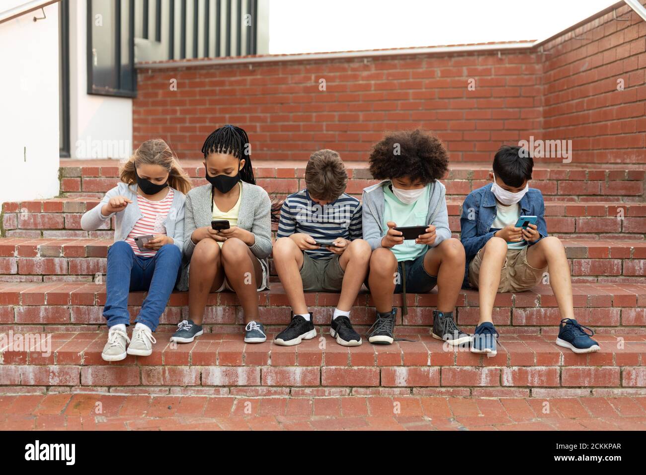 Groupe d'enfants portant un masque facial avec un smartphone lorsqu'ils sont assis sur les escaliers ensemble Banque D'Images