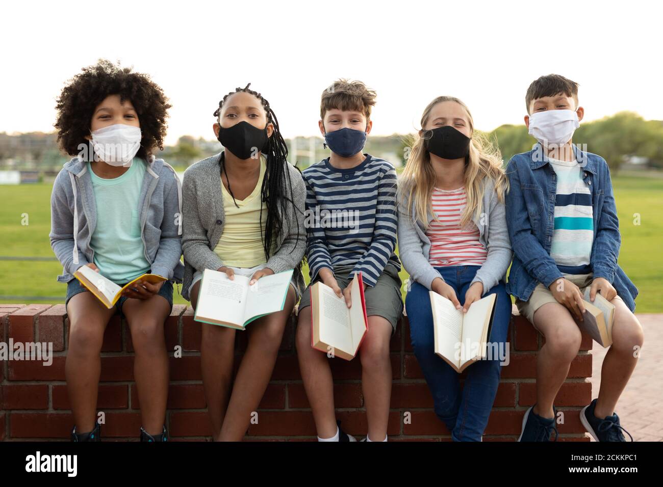 Portrait d'un groupe d'enfants portant un masque facial avec des livres assis sur un mur de briques Banque D'Images