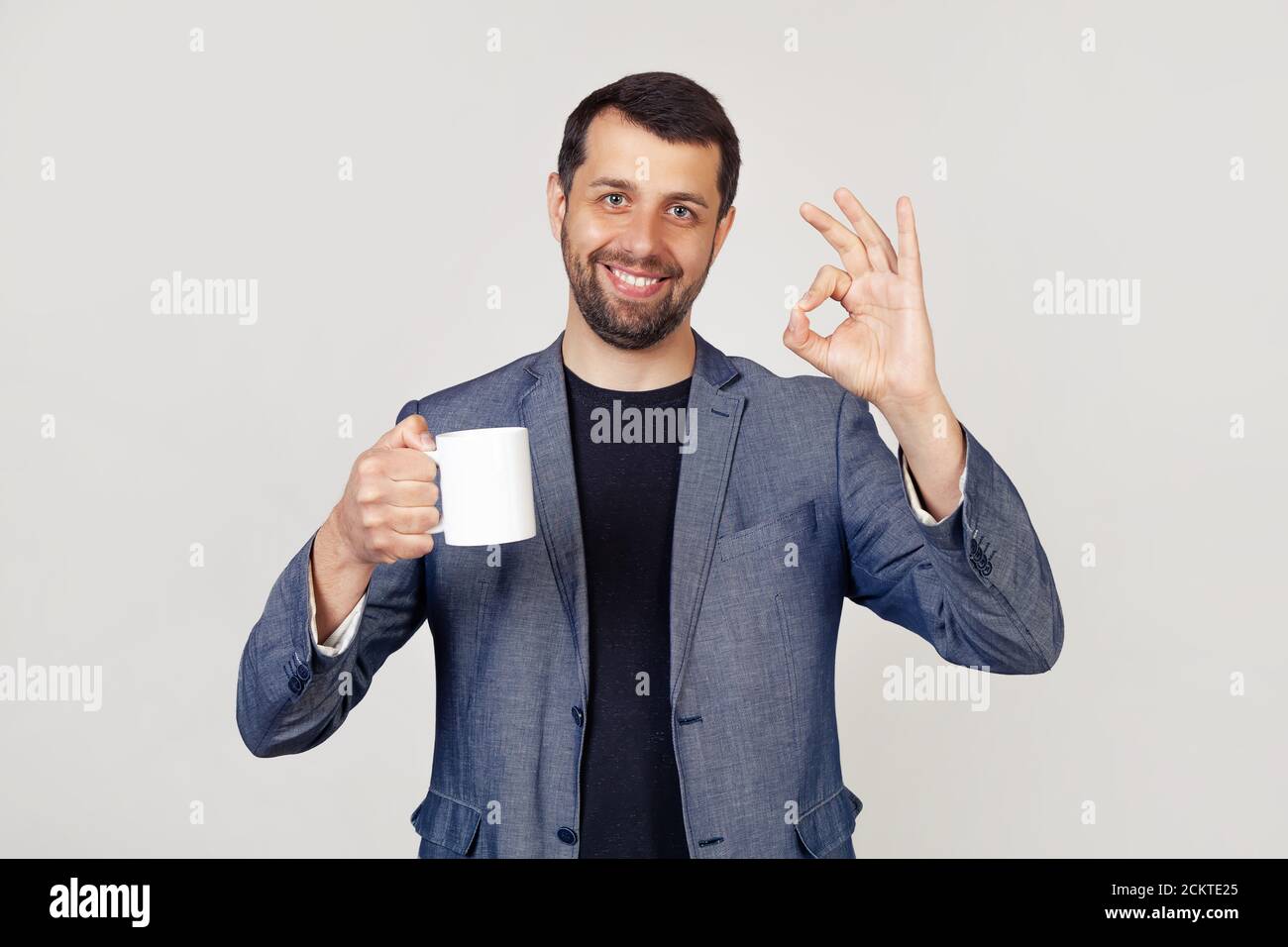 Jeune homme d'affaires avec un sourire, un homme avec une barbe dans une veste, tenant une tasse de café et montrant le geste tout est ok, un homme avec un visage heureux. Portrait d'un homme sur fond gris. Banque D'Images